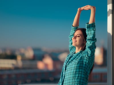 Junge Frau mit kariertem Hemd streckt sich in der Sonne. Im Hintergrund ist die Skyline einer Stadt zu sehen.