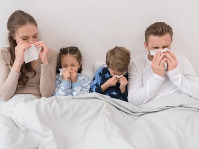 Familie ist zusammen im Bett. Alle sind krank und schnäuzen in ein Taschentuch.