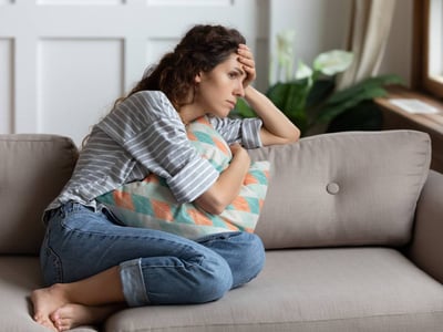 Frau sitzt auf dem Sofa und hält sich die Stirn mit einer Hand und ein Kissen mit der anderen Hand