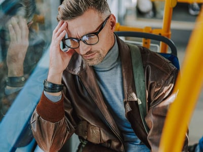 Ein Mann mit Brille sitzt im Bus und hält sich mit schmerzverzerrtem Gesicht den Kopf.