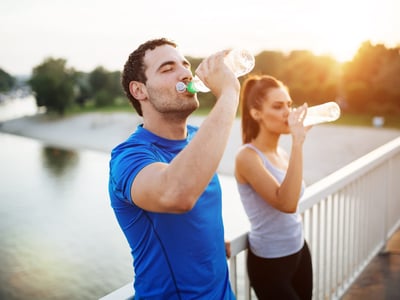 Es ist ein junges Paar in Sport-Outfits zu sehen, beide trinken Wasser.
