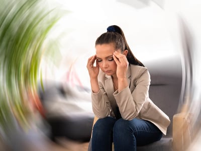 Eine Frau sitzt auf einem Sofa und hält ihren Kopf in den Händen. Ein kreisförmig verschwommener Hintergrund symbolisiert Schwindel.