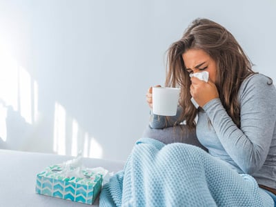 Eine erkältete Frau hält eine Teetasse in der einen Hand und putzt sich mit der andere Hand die Nase.
