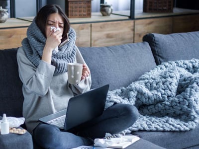 Junge Frau sitzt auf der Couch und putzt sich die Nase. Auf ihrem Schoß steht ein Laptop.