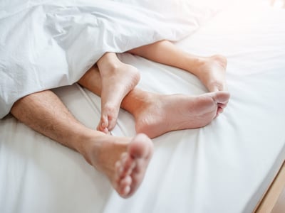 In einem weiß bezogenen Bett gucken die Füße von einem Mann und einer Frau unten aus der Bettdecke.