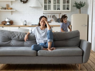 Eine Frau sitzt genervt auf dem Sofa, während Kinder um sie herumtoben.