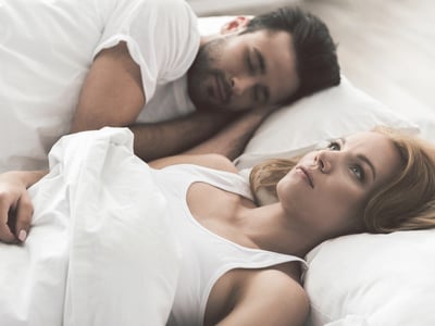 EIne junge Frau liegt wach im Bett und starrt nach oben, daneben schläft ein Mann.