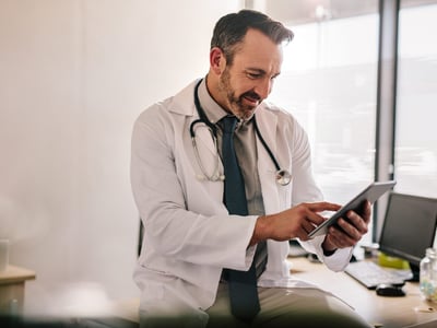 Arzt sitzt auf der Ecke eines Schreibtischs und nutzt ein Tablet.