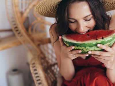 Es ist eine junge Frau mit dunklen Haaren zu sehen, die einen Strohhut trägt und in ein großes Stück Melone beist.