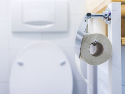 Eine Rolle Toilettenpapier hängt im Badezimmer. Im Hintergrund ist eine Toilette zu sehen.