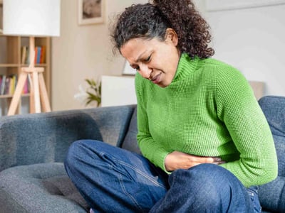 Frau sitzt zusammengekrümmt auf dem Sofa und hält sich den schmerzenden Bauch.