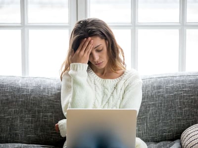 Eine Frau sitzt mit Laptop auf dem Sofa und hat Kopfschmerzen.
