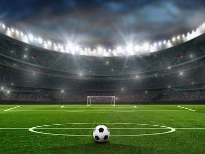 Fussball liegt im Stadion vor der Mittellinie. Im Hintergrund Flutlicht und ein leeres Tor.