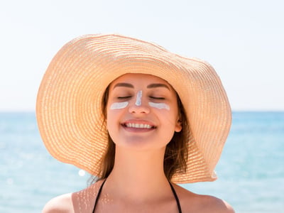 Junge Frau am Meer, die einen Sonnenhut trägt und Sonnencreme im Gesicht hat.