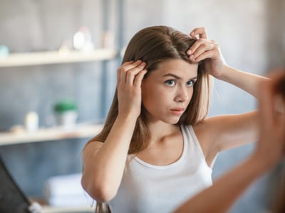 Junge Frau betrachtet sich im Spiegel, sie prüft ihren Haaransatz auf Haarausfall.