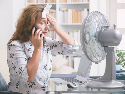 Frau am Telefon sitzt vor dem Ventilator und tupft sich die Stirn mit einem Tuch.