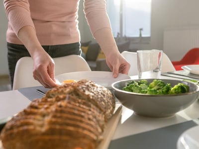 Im Bild ist ein gedeckter Tisch zu sehen, darauf aufgeschnittenes Brot und Broccoli. Eine Frau stellt einen Teller auf den Tisch.