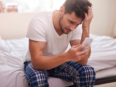 Mann sitzt auf Bettkante und schaut traurig und verzweifelt auf sein Handy.
