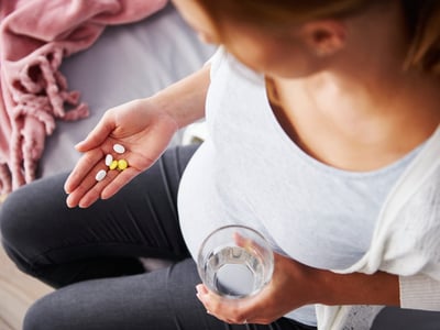 Eine schwangere Frau hält Nahrungsergänzungsmittel in einer Hand und ein Glas Wasser in der anderen.