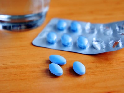 Blaue Tabletten in einem Blister liegen auf einem Holztisch, im Hintergrund steht ein Glas Wasser.