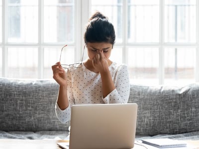 Eine junge Frau sitzt auf der Couch, vor ihr steht ein Laptop. Sie hat ihre Brille abgenommen, schließt die Augen und wirkt gestresst.