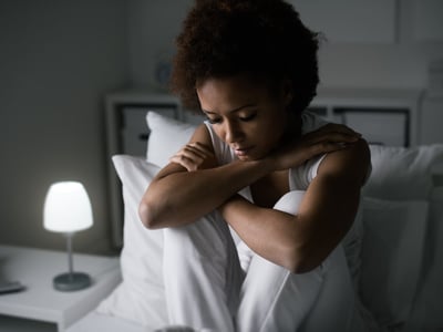 Eine junge Frau sitzt niedergeschlagen auf einem Bett.