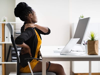 Eine Frau sitzt im Büro am Schreibtisch und hält sich den Schmerzenden Rücken.