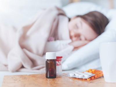 Schlaftabletten stehen auf einem Nachttisch, im Hintergrund schläft eine Frau im Bett.