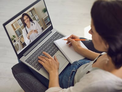 Eine Frau sitzt am Laptop und macht sich während der Online-Sprechstunde mit einer Ärztin Notizen.