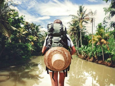 Frau macht einen Abenteuerurlaub. Sie steht mit Rucksack und Strohhut an einem Fluss.