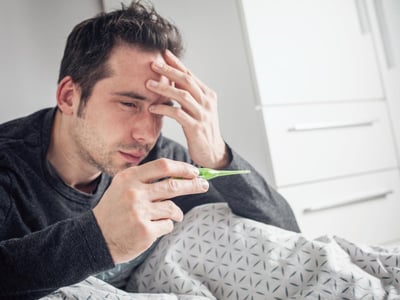 Ein Mann ist krank und sitzt im Bett. Er hat ein Fieberthermometer in der Hand.