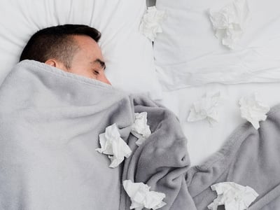 Ein Mann liegt im Bett. Er ist bis zur Nase zugedeckt und es liegen viele benutzte Taschentücher um ihn herum.