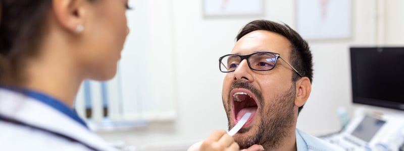 Eine Ärztin drückt bei ihrem Patienten die Zunge mit einem Spatel runter um Hals und Rachen zu untersuchen.