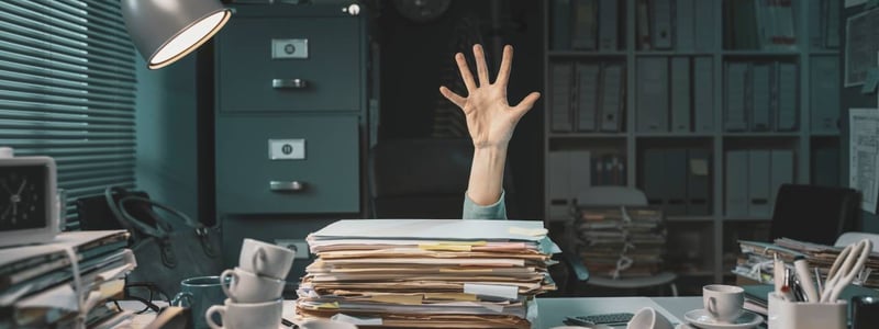Ein voller Schreibtisch steht in einem Büro. In der Mitte des Bildes ist eine ausgestreckte Hand. Es ist ein Symbolbild für jemanden, der in Arbeit ertrinkt.