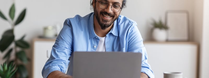 Junger Mann mit dunklen, längeren Haaren und hellblauen Hemd sitzt vor Laptop.