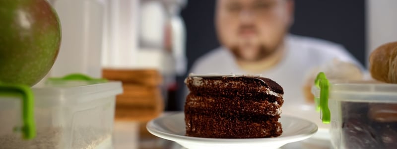 Es ist ein Mann zu sehen, der die Kühlschranktür öffnet. Im Fokus des Bildes ist ein Schokokuchen.