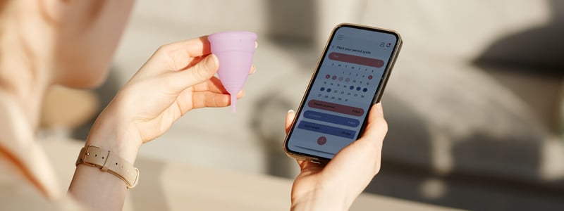 Frau hält Menstruationstasse in der linken Hand und Handy mit Zykluskalender in der rechten.