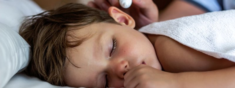 Kind mit Ohrenschmerzen liegt im Bett und bekommt Ohrentropfen verabreicht.