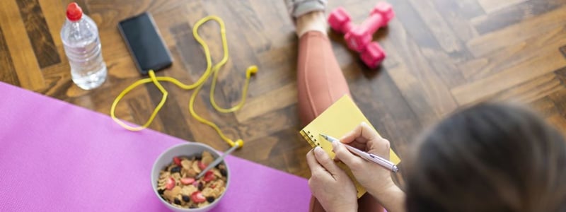 Kombination aus Ernährung und Sport um abzunehmen. Junge Frau schreibt in ein Notizbuch, sitzt auf Yogamatte und hat gesundes Müsli und Hanteln vor sich.