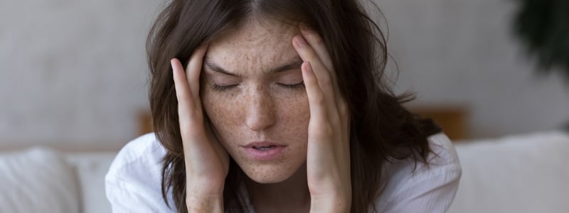 Junge Frau mit Sommersprossen hat Kopfschmerzen und hält sich den Kopf.