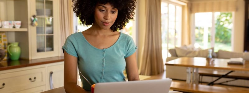 Junge Frau in blauen T-Shirt sitzt in der Küche vor ihrem Laptop.