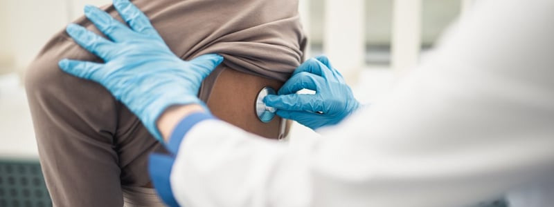 Ein Arzt hört mit dem Stethoskop am Rücken einer Patientin die Lunge ab.