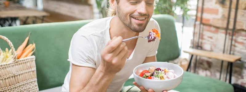 Auf dem Bild ist ein junger Mann zu sehen, der auf einem grünem Sofa sitzt und einen Salat isst.