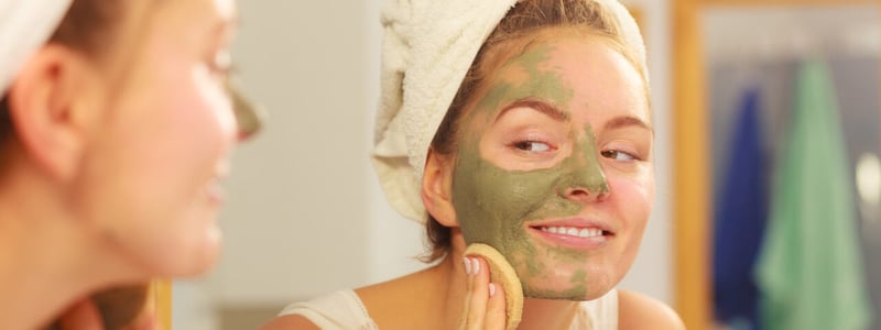 Junge Frau vor dem Spiegel im Badezimmer mit einer Gesichtsmaske aus Heilerde.