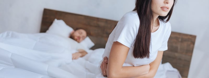 Frau sitzt auf Bettkante und hält sich vor Schmerzen den Unterleib.
