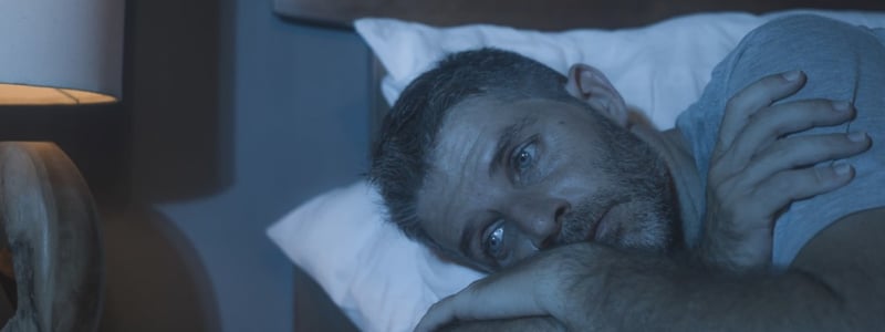 Ein Mann liegt in einem abgedunkelten Raum schlaflos im Bett.