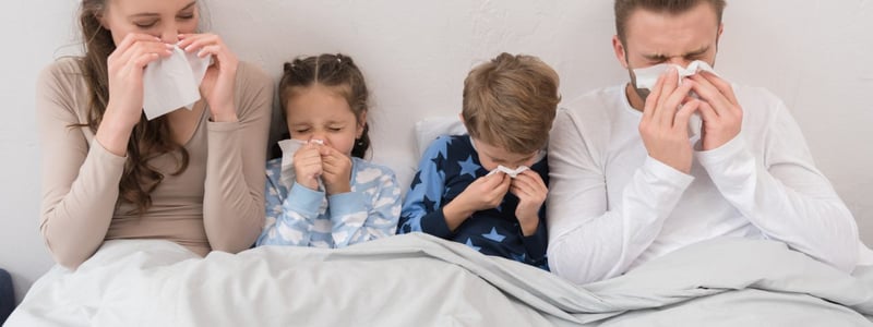 Familie ist zusammen im Bett. Alle sind krank und schnäuzen in ein Taschentuch.