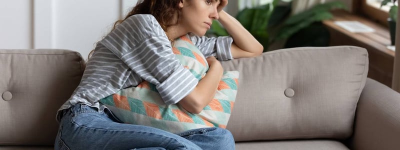Frau sitzt auf dem Sofa und hält sich die Stirn mit einer Hand und ein Kissen mit der anderen Hand