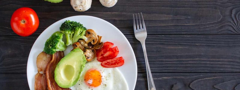 Es ist ein Teller mit ketogenen Lebensmitteln zu sehen, wie Avocado, Tomate, Pilzen, Broccoli, Speck und Ei.