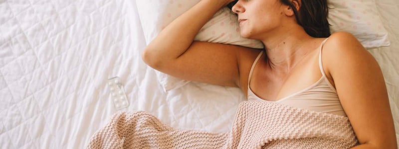 Frau liegt mit Schmerzen im Bett und hält sich den Bauch, neben ihr liegt eine Packung Schmerztabletten.